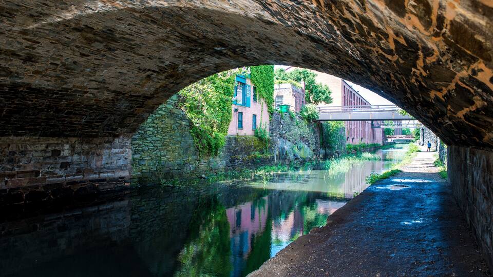 A stone bridge arching over a calm canal, with buildings and greenery in the background.