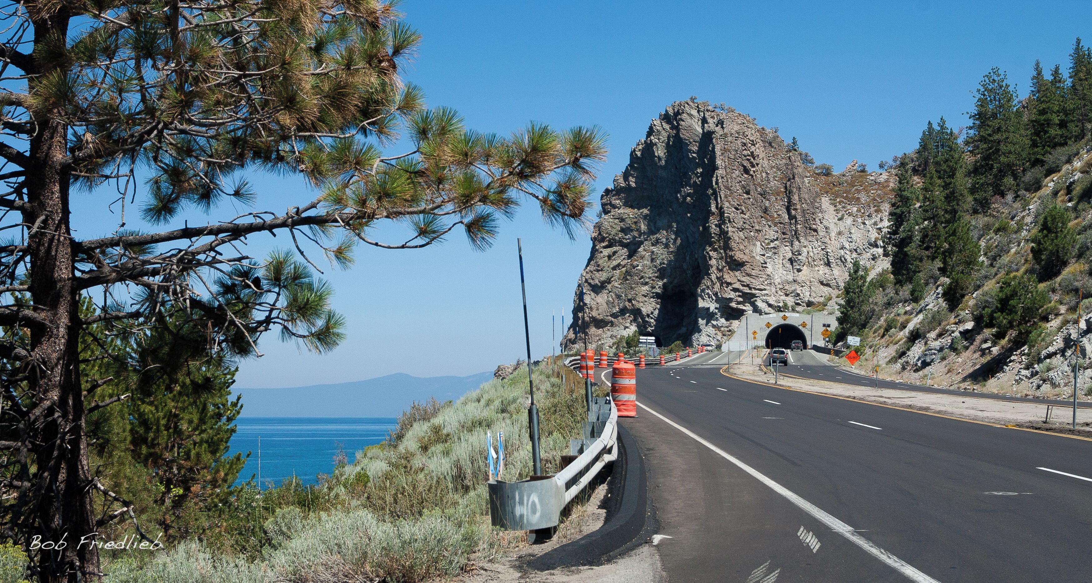 A coastal road with a rocky cliff, pine trees, and a clear blue sky.