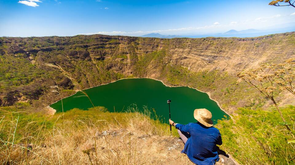 A person taking a photo of a green lake surrounded by hills.