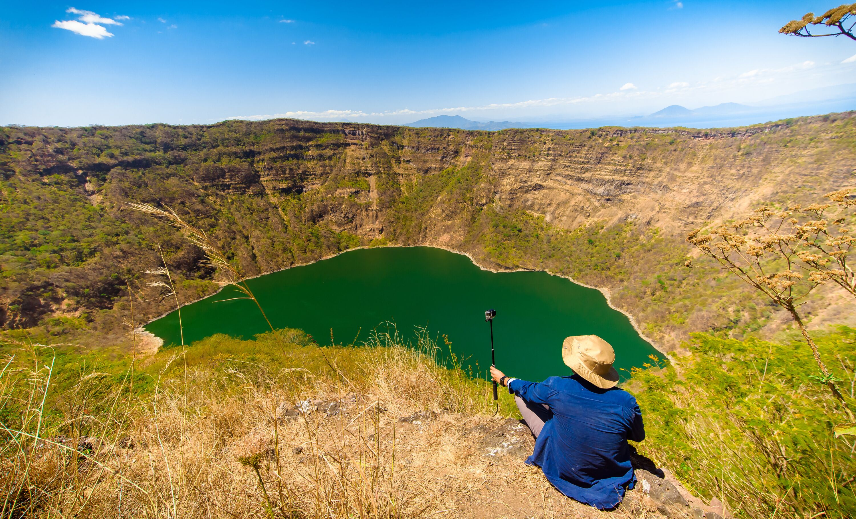 A person taking a photo of a green lake surrounded by hills.