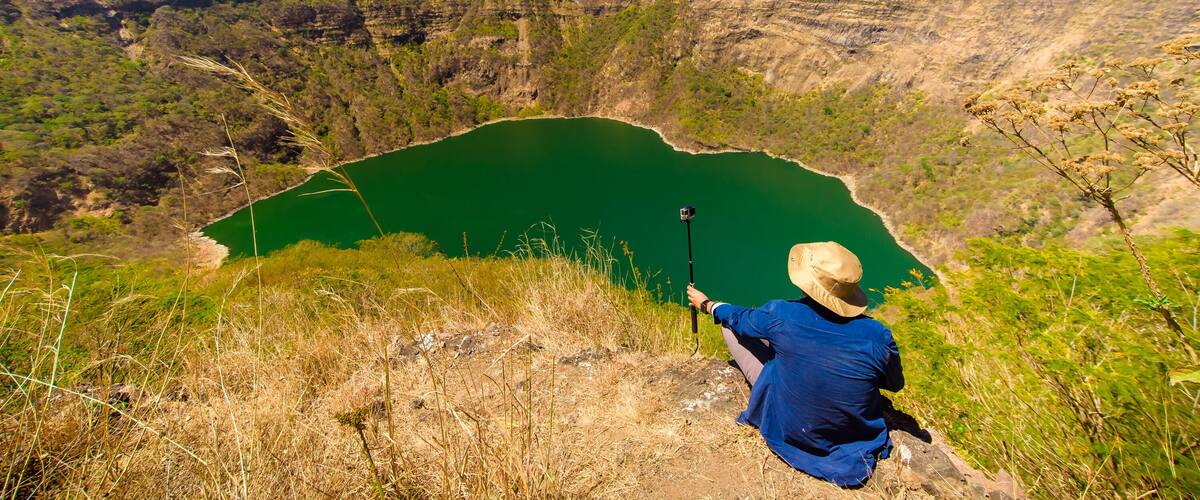 A person taking a photo of a green lake surrounded by hills.