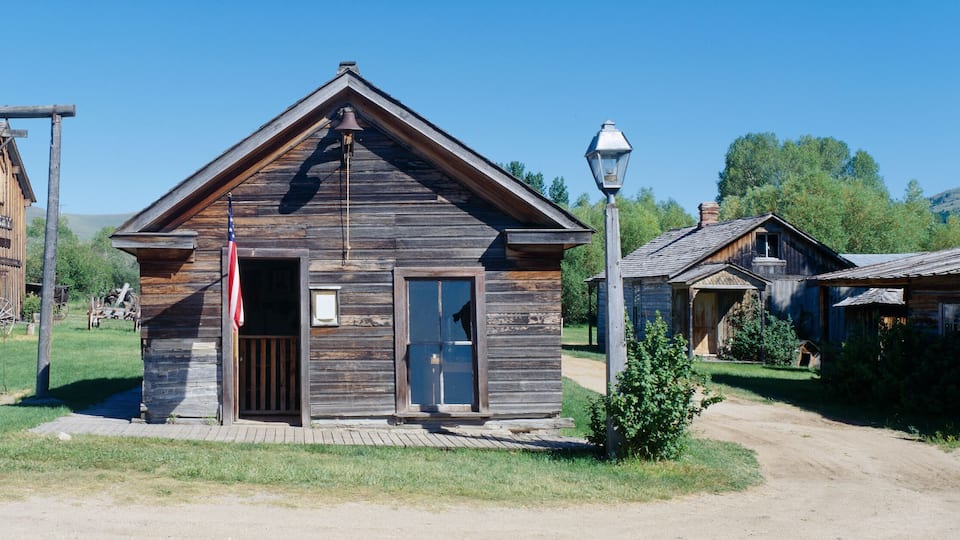 A rustic wooden building with a bell tower, surrounded by other wooden structures in a rural setting.