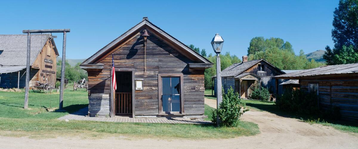 A rustic wooden building with a bell tower, surrounded by other wooden structures in a rural setting.
