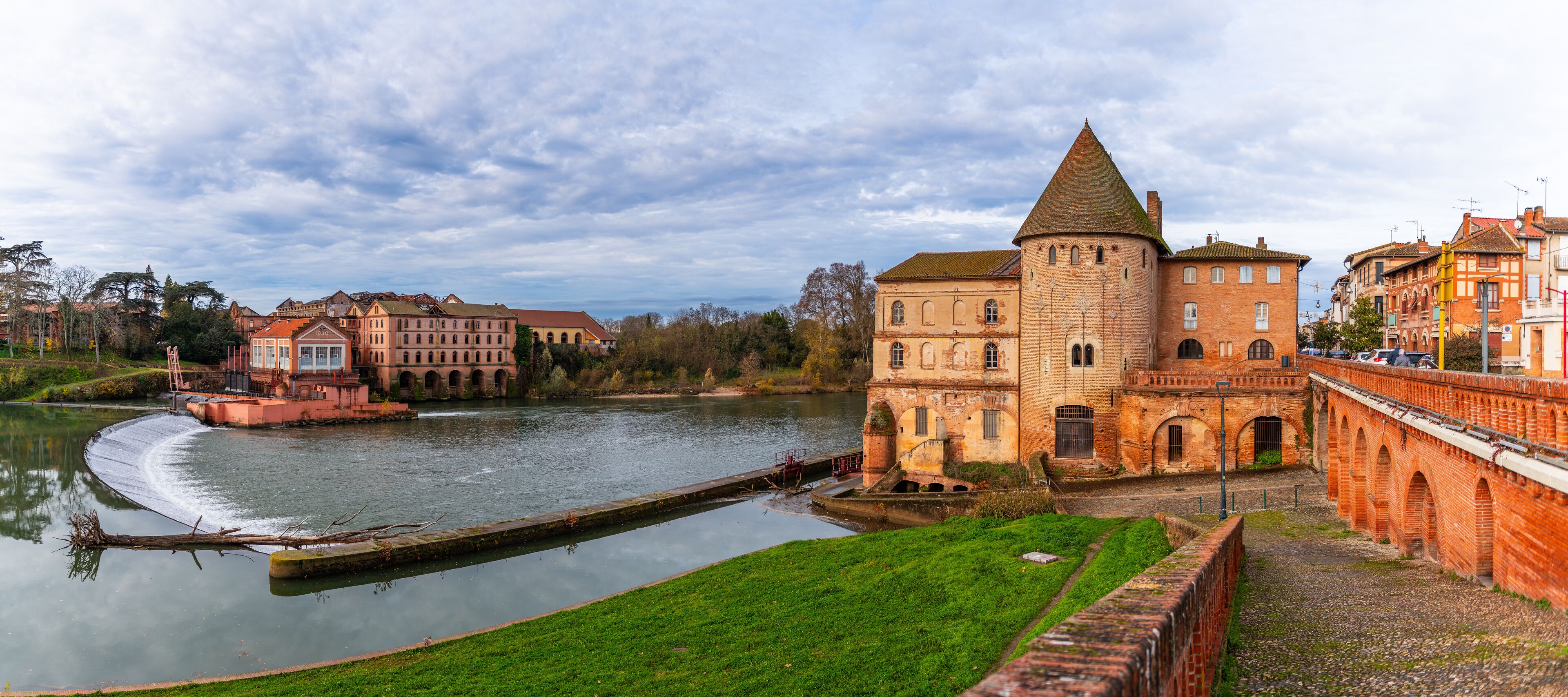A historic building with a tower by a river, surrounded by other buildings and greenery.