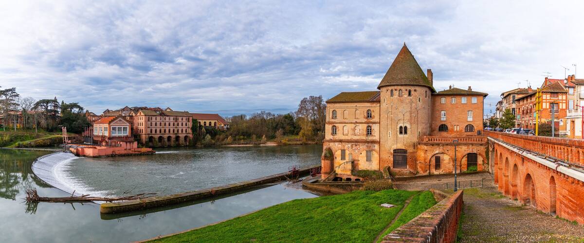 A historic building with a tower by a river, surrounded by other buildings and greenery.