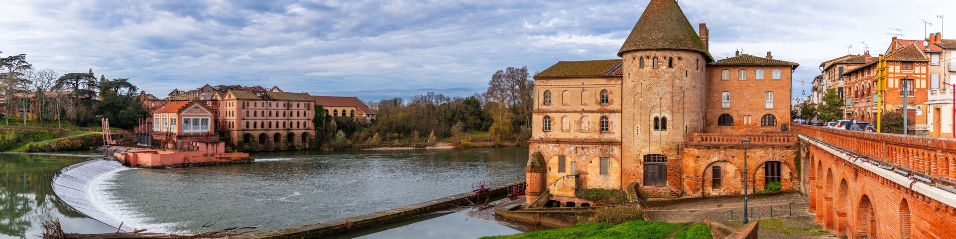A historic building with a tower by a river, surrounded by other buildings and greenery.