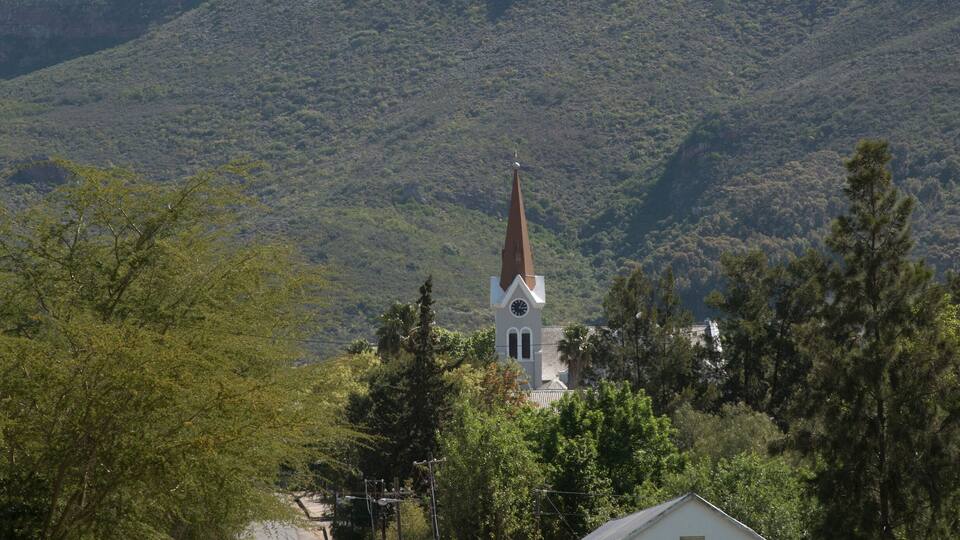 A road leading to a church with a steeple, surrounded by trees and a mountain in the background.