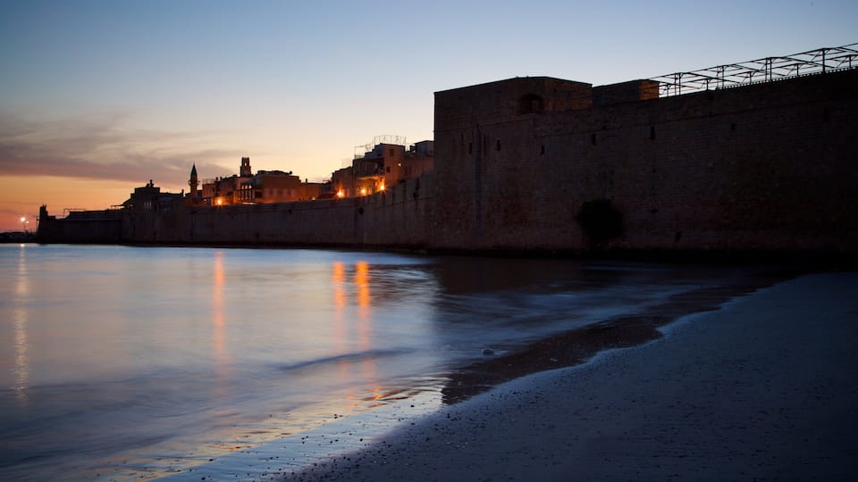 Ein Küstenstadtbild in der Dämmerung mit einer markanten Steinmauer und beleuchteten Gebäuden, die sich im ruhigen Meer spiegeln.
