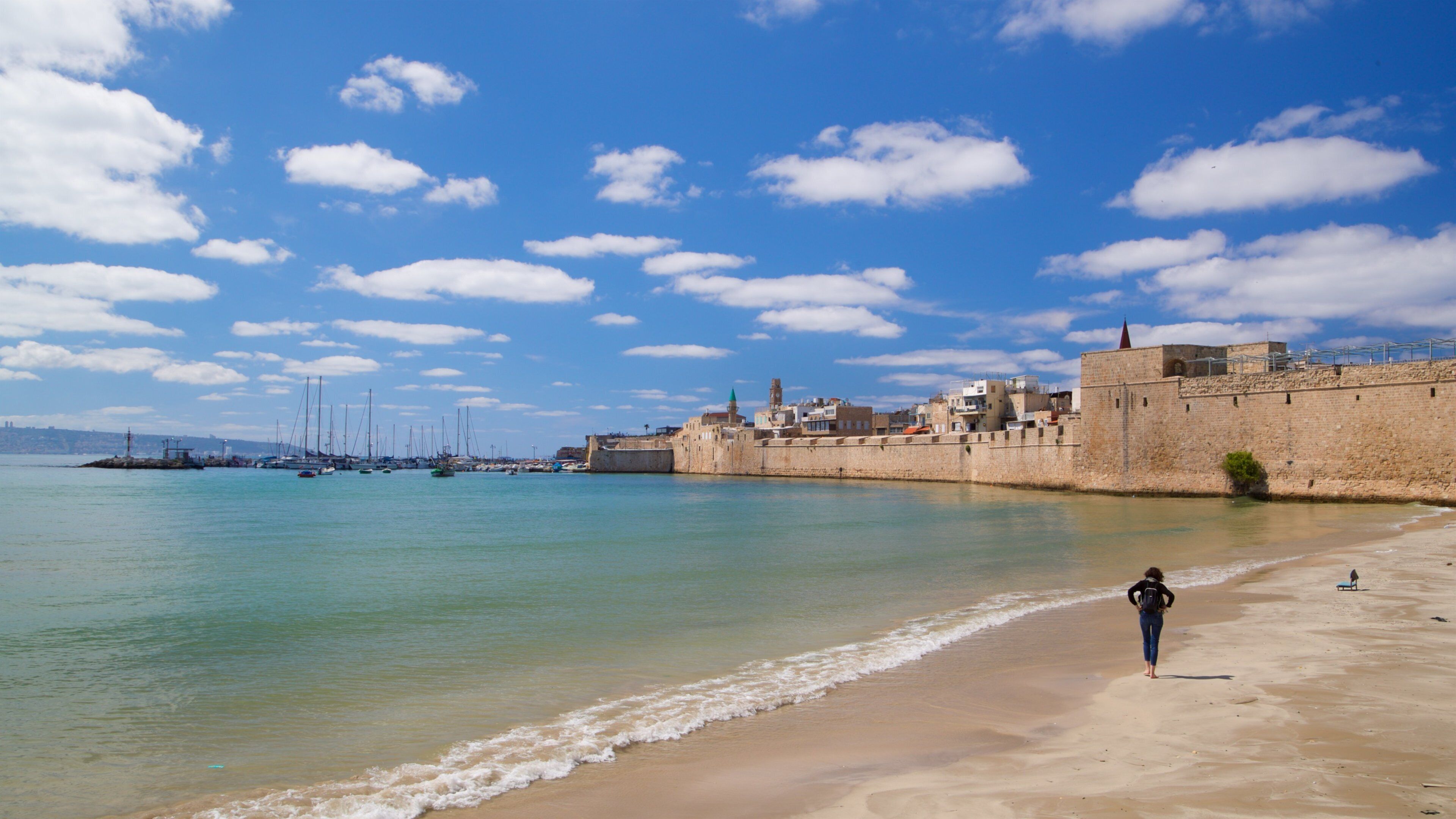 A coastal town with a sandy beach, a historic stone wall, and sailboats in the harbor.