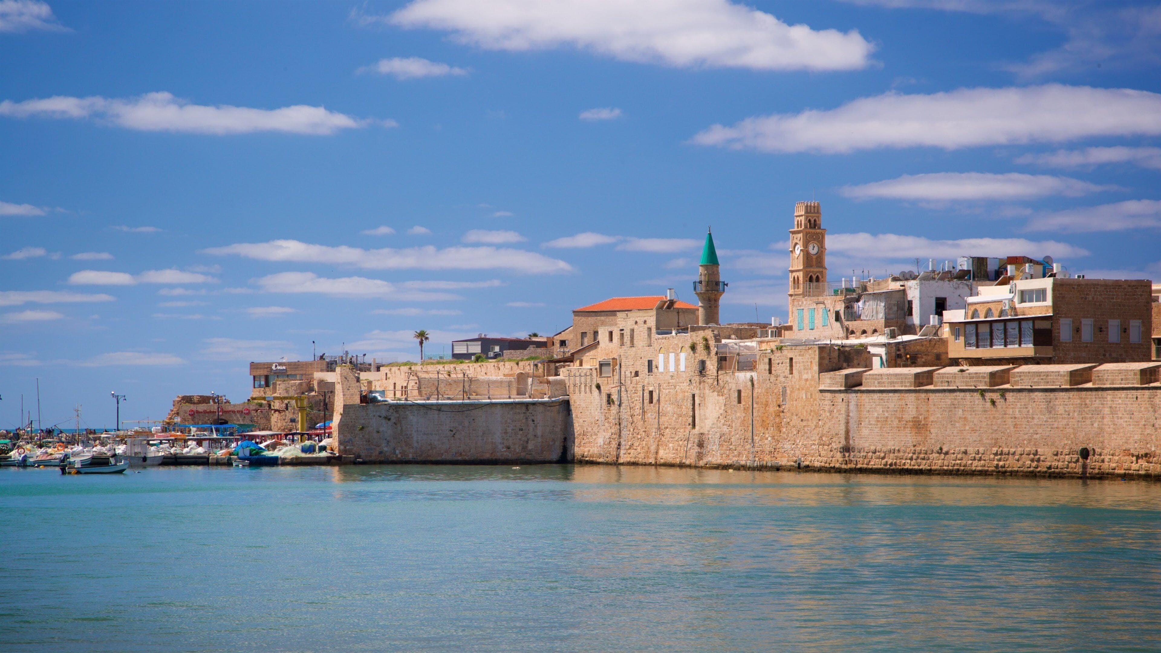 A coastal cityscape with historic buildings, a prominent clock tower, and a calm harbor with boats.