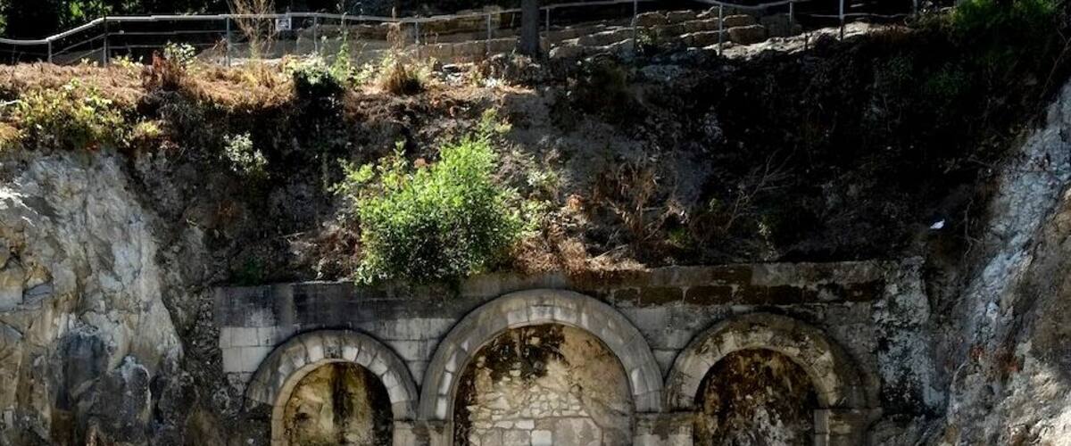 An ancient stone structure with three arched doorways and a central door, surrounded by rocky terrain and sparse vegetation.