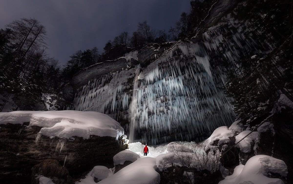 A person in a red jacket standing in front of a waterfall with icicles.