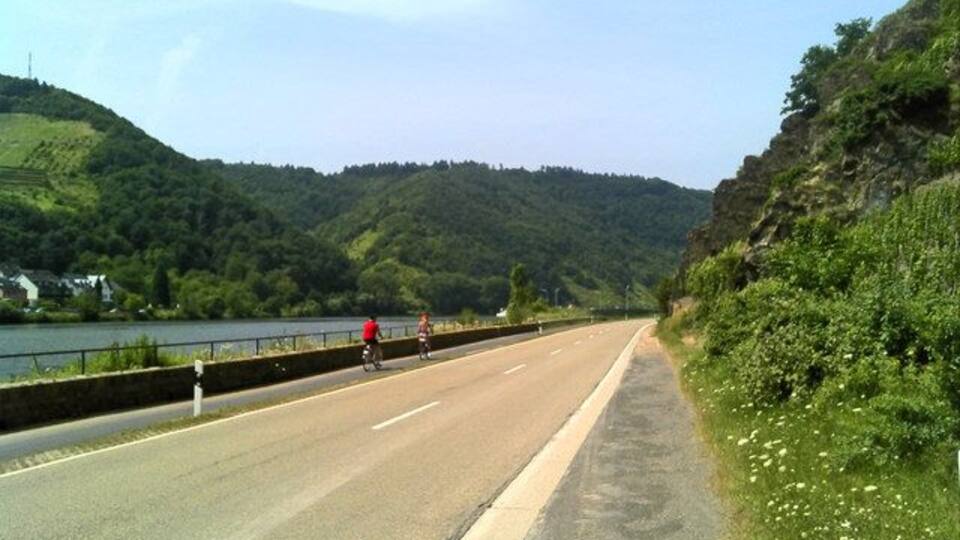A road with cyclists, a river, and hills in the background.