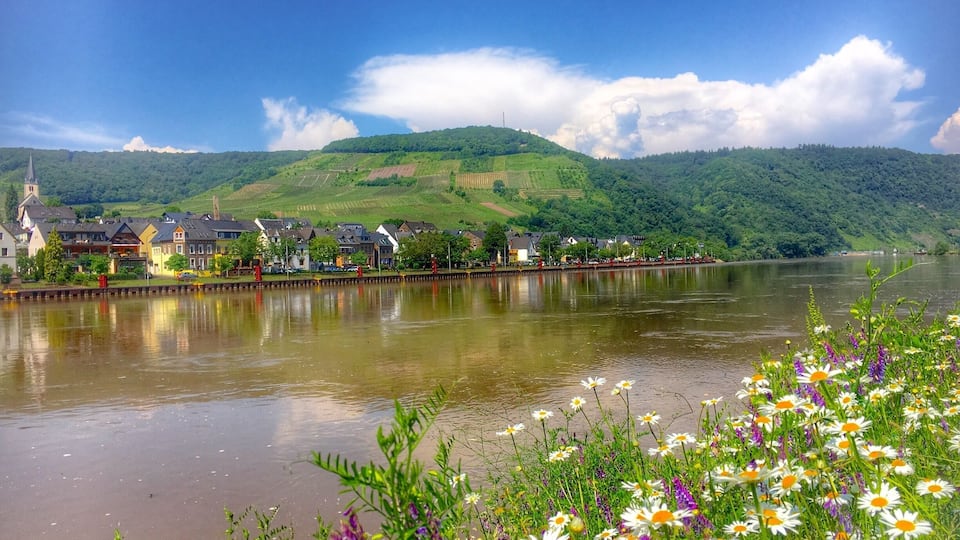 A river with a town on the opposite bank, surrounded by green hills and a clear sky.