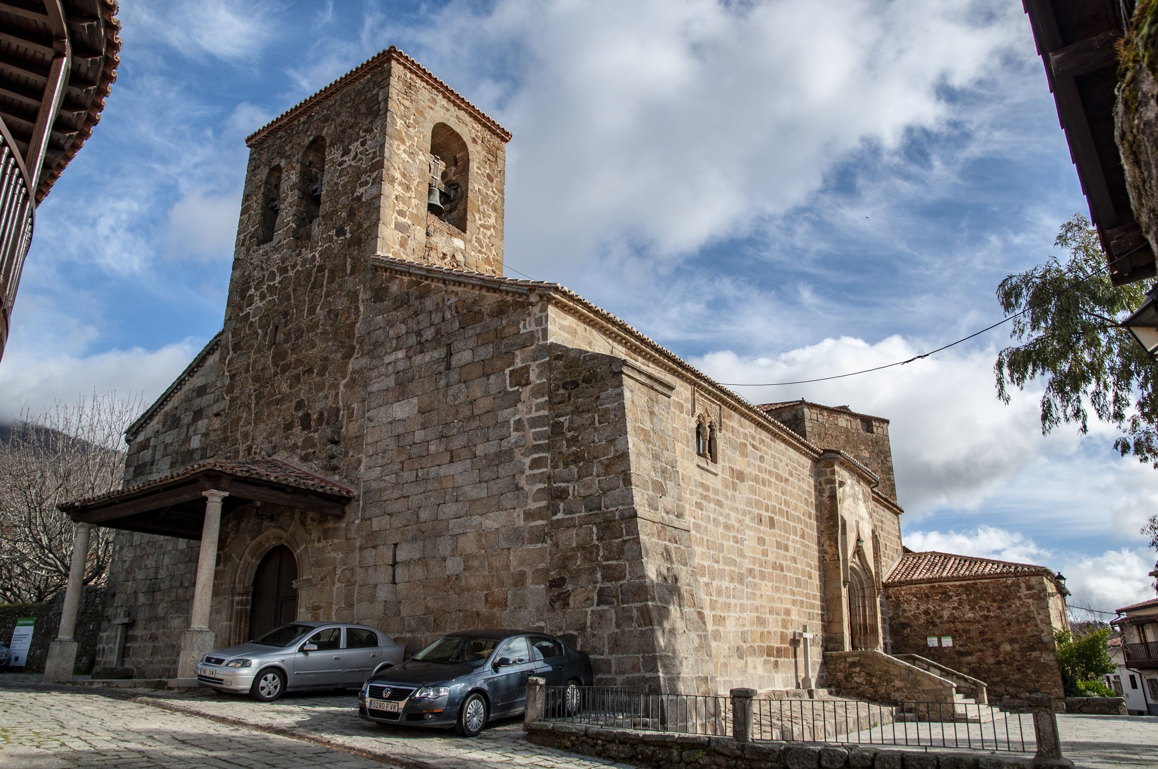 A historic stone church with a bell tower, parked cars, and a cobblestone street.
