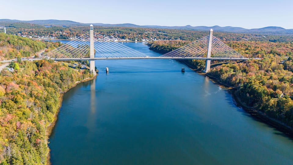 A suspension bridge over a river with surrounding autumn foliage and distant hills.