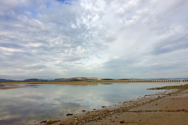A calm river with a bridge, rocky shoreline, and distant mountains under a cloudy sky.