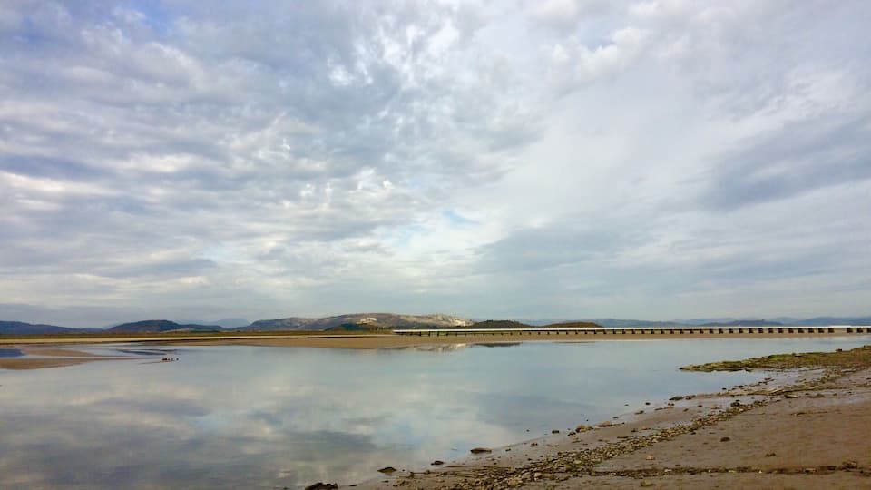 A calm river with a bridge, rocky shoreline, and distant mountains under a cloudy sky.