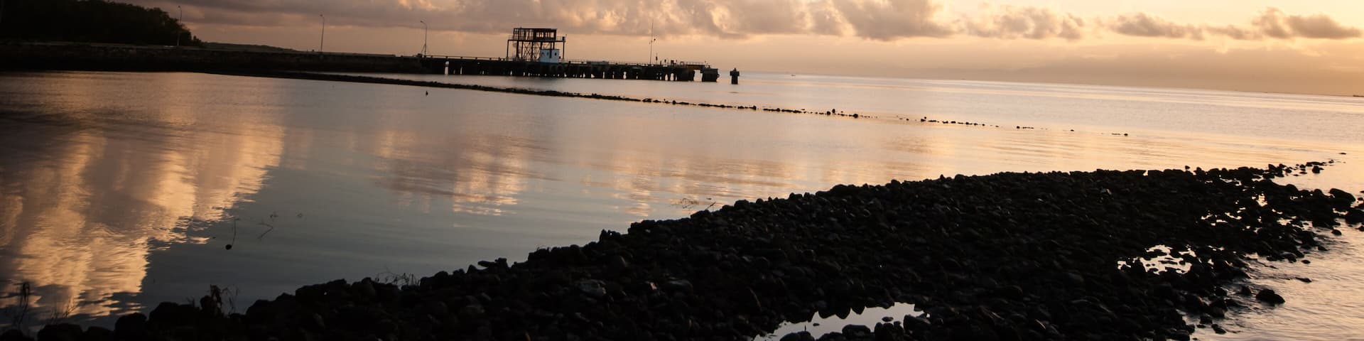 A calm lake with a pier extending into the water, surrounded by a rocky shoreline and a backdrop of clouds.