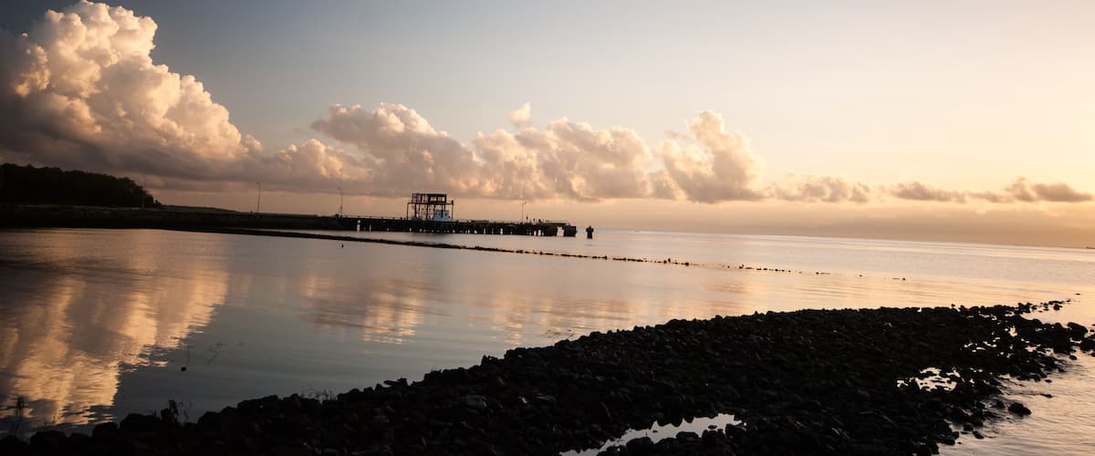 A calm lake with a pier extending into the water, surrounded by a rocky shoreline and a backdrop of clouds.