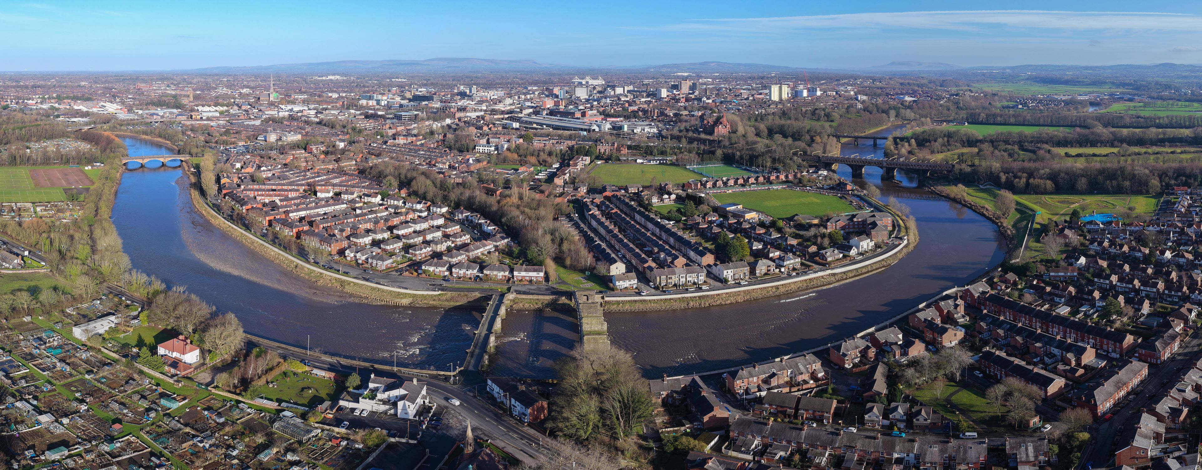 An aerial view of a river winding through a residential area with a bridge and industrial buildings in the distance.