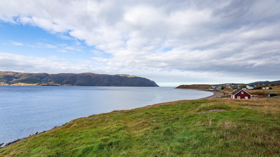 A coastal landscape with a red barn, grassy fields, and a calm sea under a cloudy sky.