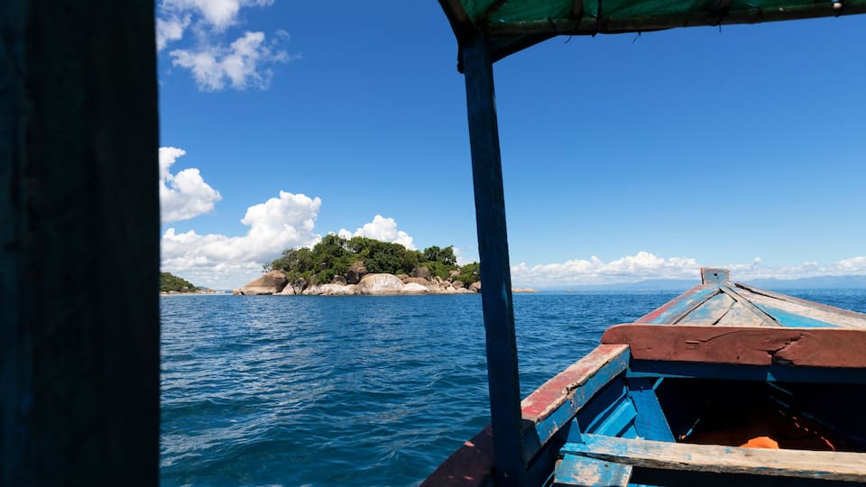 A boat with a blue and red hull on a calm sea, a rocky island with greenery in the distance, and a clear blue sky with clouds.