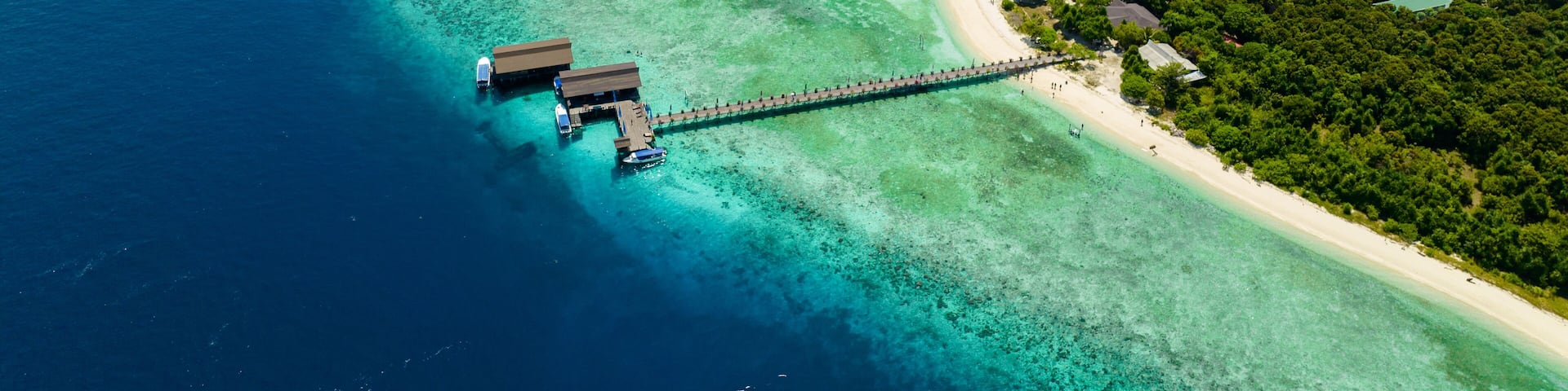 An aerial view of a tropical island with clear turquoise waters, a sandy beach, and a wooden pier connecting to overwater bungalows.