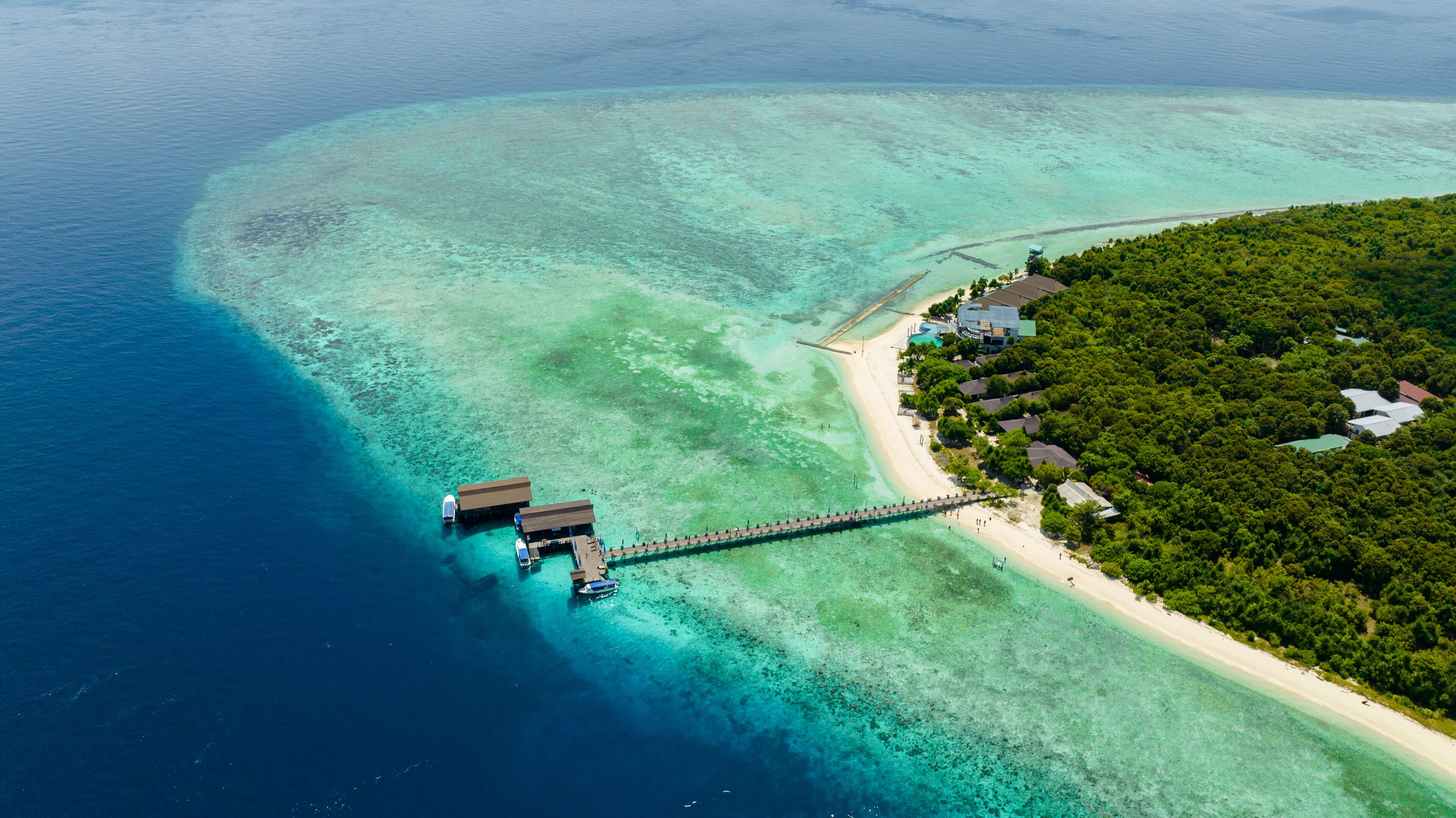 An aerial view of a tropical island with clear turquoise waters, a sandy beach, and a wooden pier connecting to overwater bungalows.