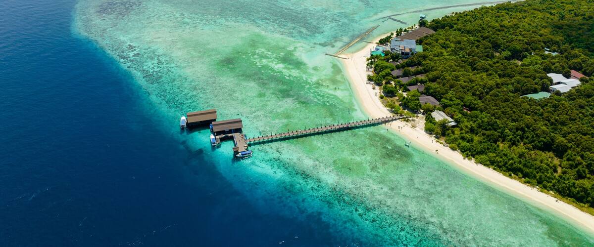An aerial view of a tropical island with clear turquoise waters, a sandy beach, and a wooden pier connecting to overwater bungalows.