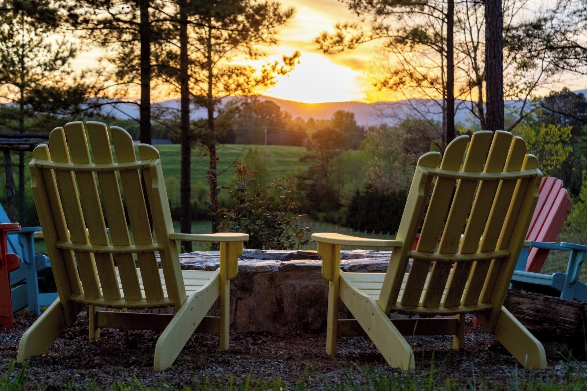 Two Adirondack chairs facing a stone fire pit with a sunset in the background.