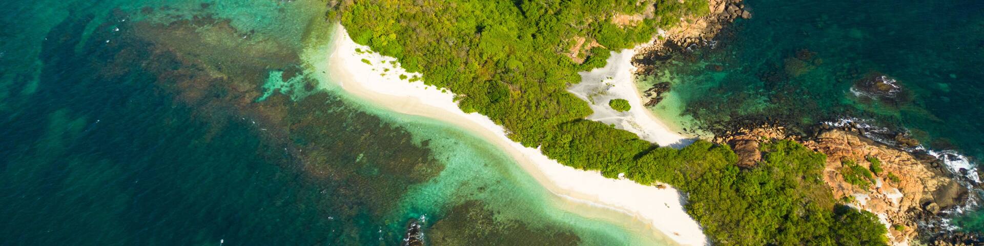 An aerial view of a tropical island with a white sandy beach, lush green vegetation, and clear turquoise waters.