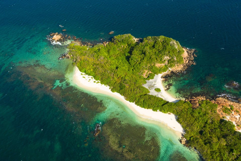 Vista aérea de una isla tropical con una playa de arena blanca, vegetación exuberante y aguas turquesas cristalinas.