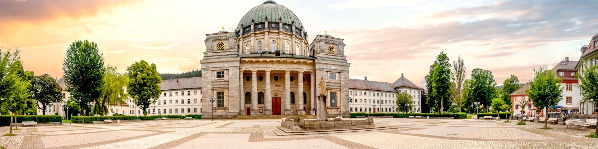 A historic building with a dome and columns, surrounded by a cobblestone square and trees.