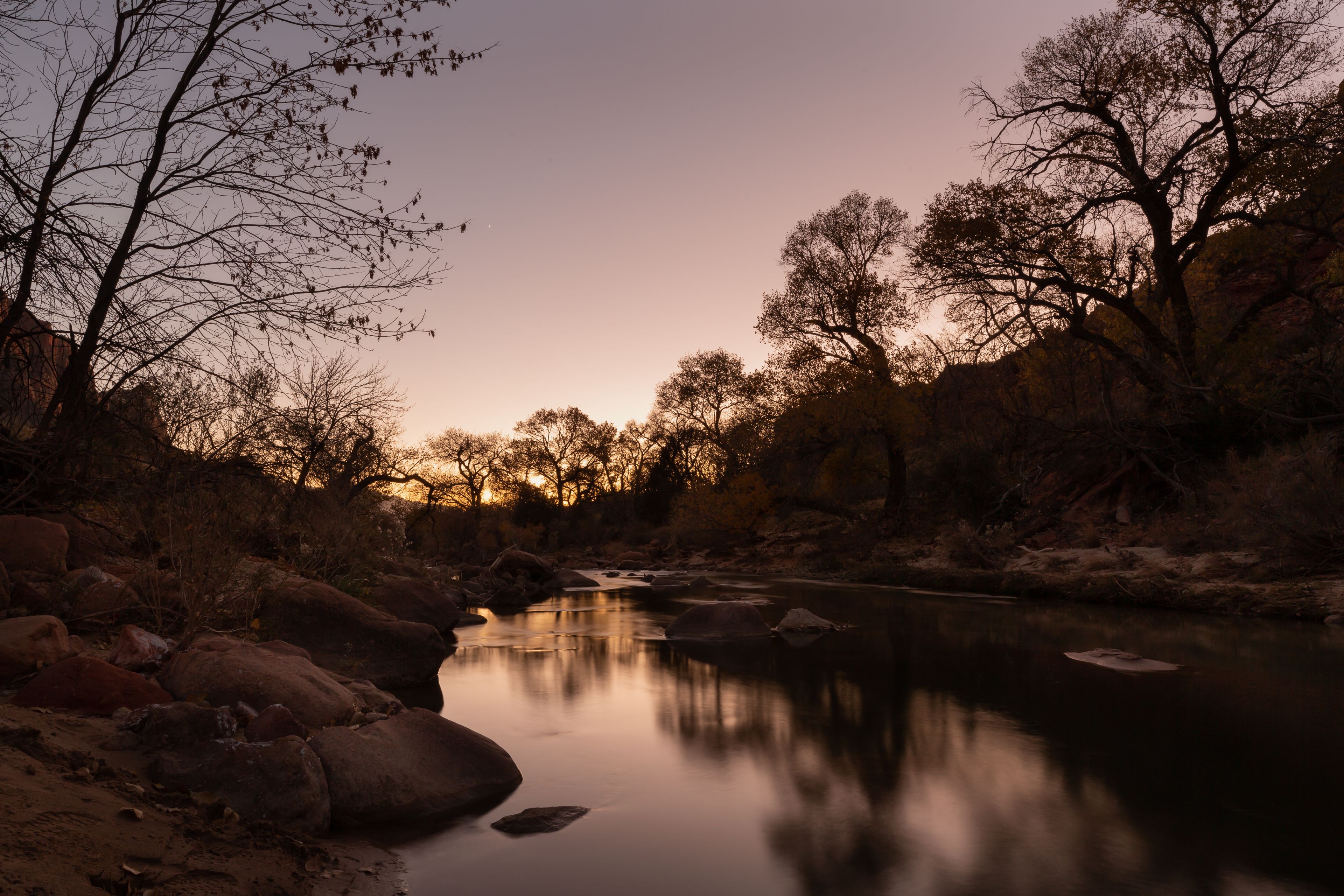 Still water in the Virgin river reflects a glowing sunset sky and winter bare trees while a single evening star begins to glow above.  