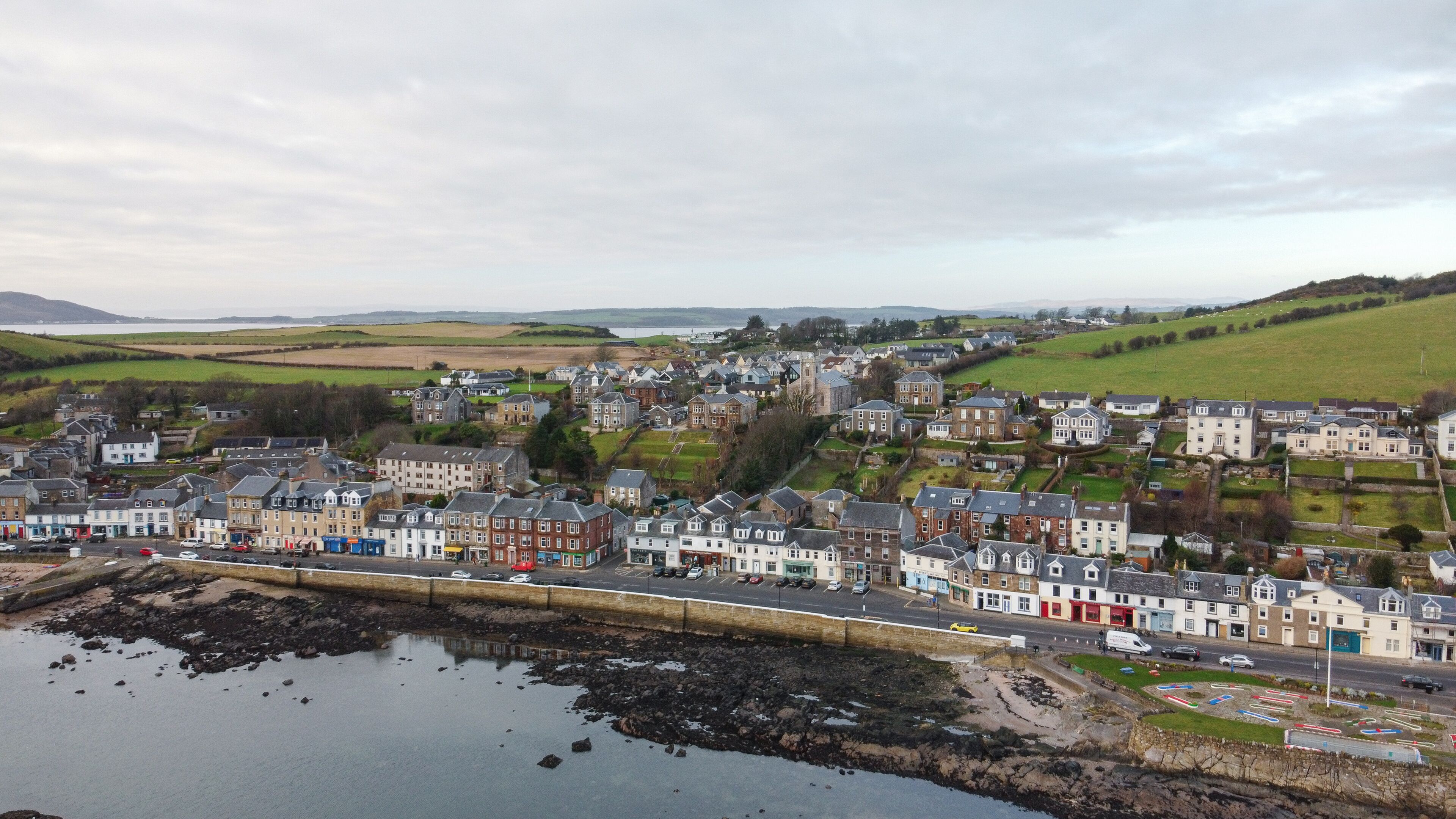 A coastal town with a mix of residential and commercial buildings, a rocky shoreline, and a large body of water.