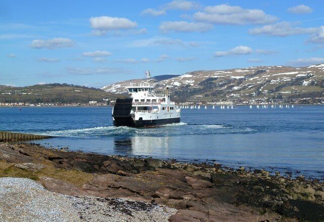 A ferry moving across a calm body of water with a rocky shoreline and a hilly landscape in the background.