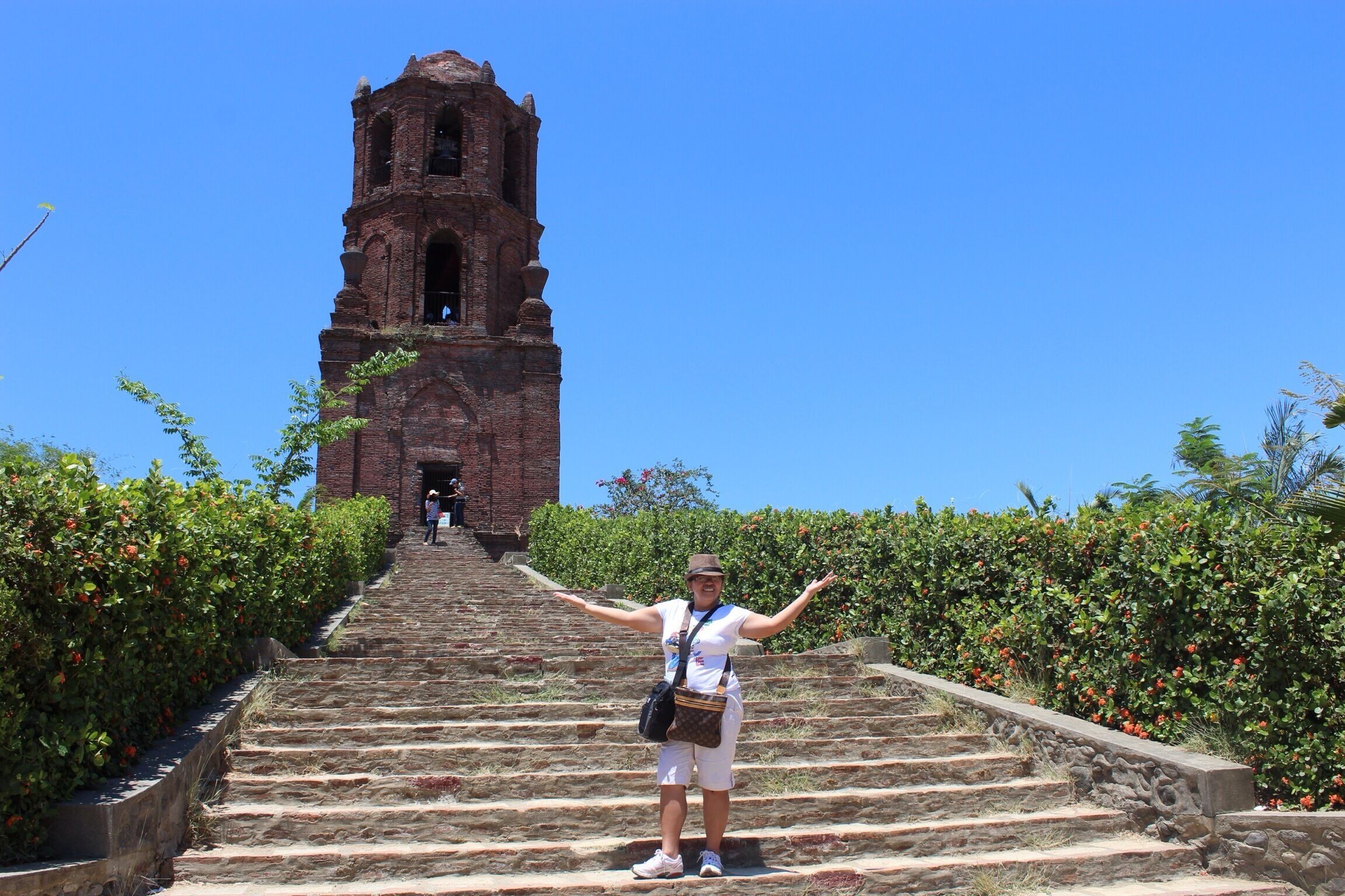 A person standing on stone steps with arms outstretched, facing a tall brick tower.