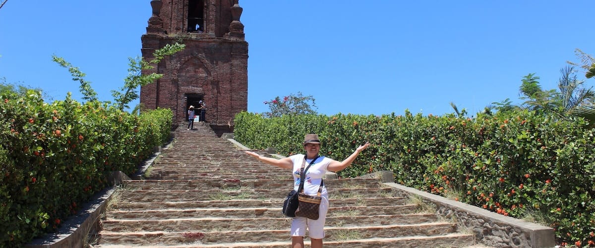 A person standing on stone steps with arms outstretched, facing a tall brick tower.