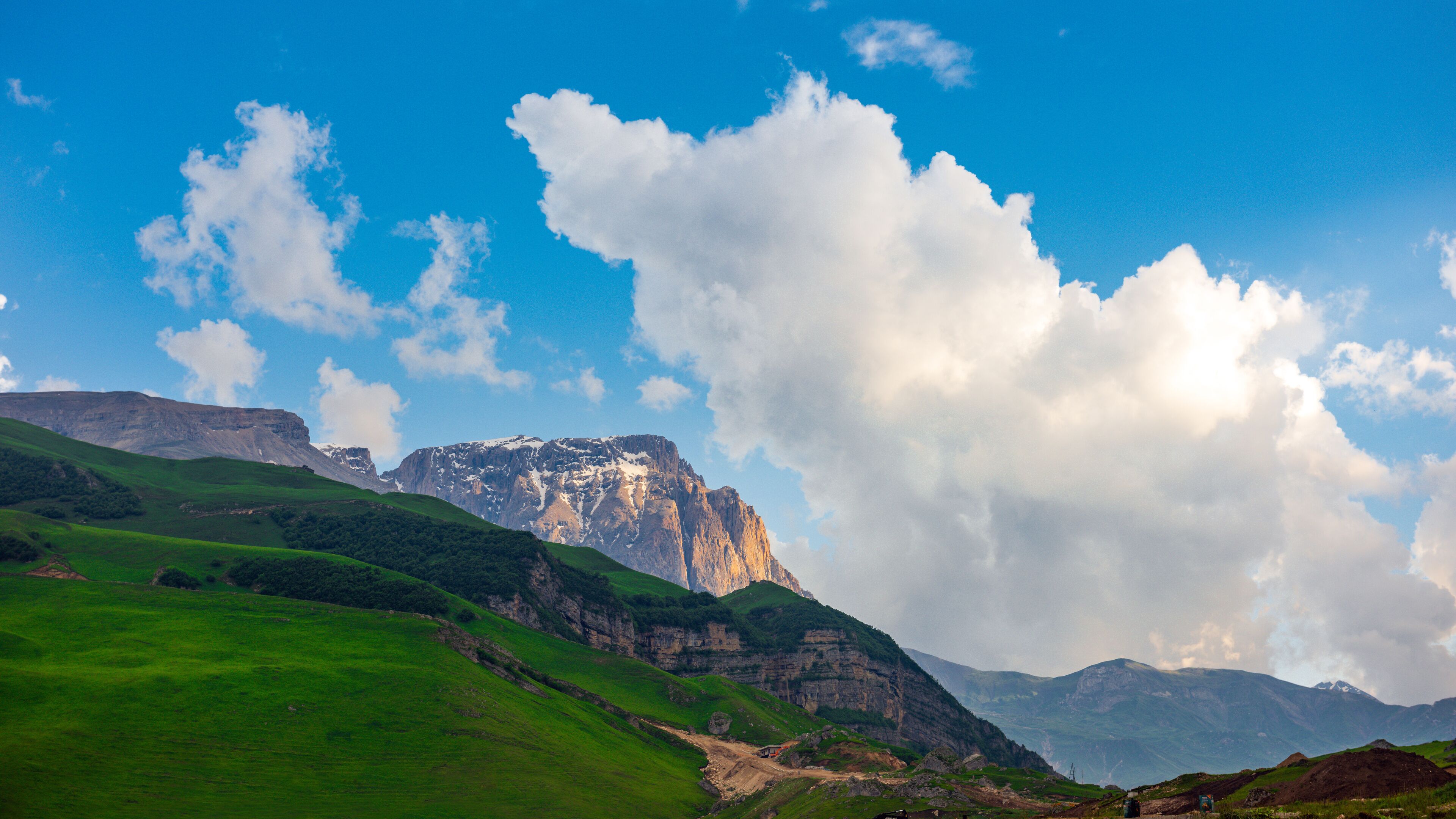 A mountainous landscape with green hills, a prominent rocky peak, and a clear blue sky with clouds.