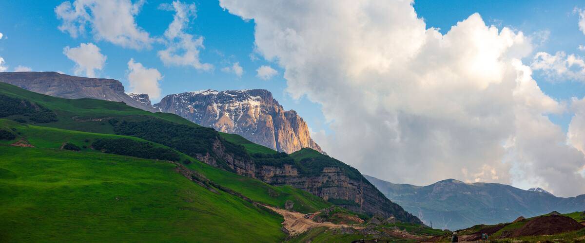 A mountainous landscape with green hills, a prominent rocky peak, and a clear blue sky with clouds.