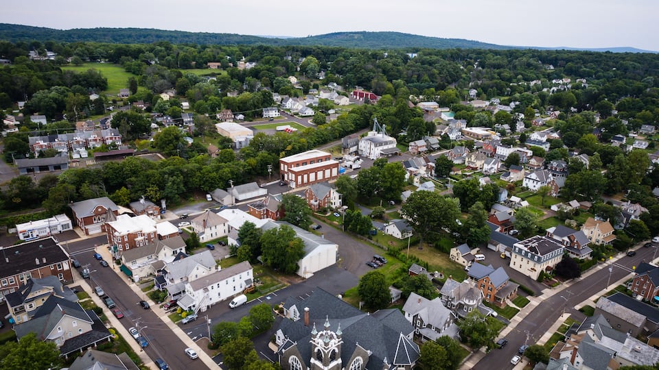 An aerial view of a small town with a mix of residential and commercial buildings, surrounded by green hills.