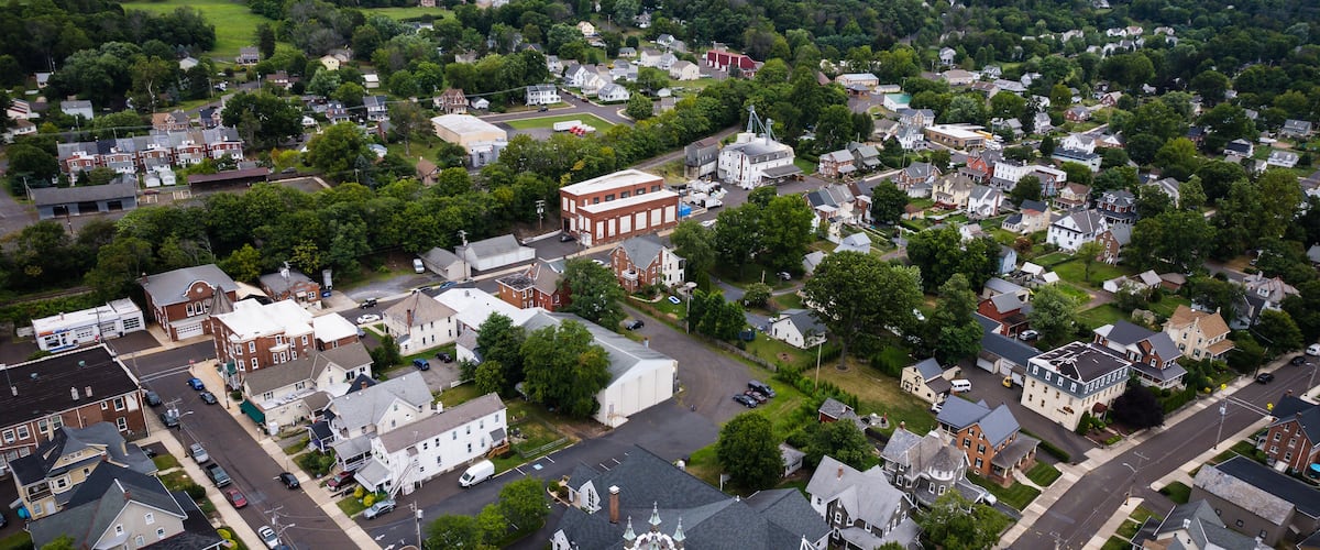 An aerial view of a small town with a mix of residential and commercial buildings, surrounded by green hills.