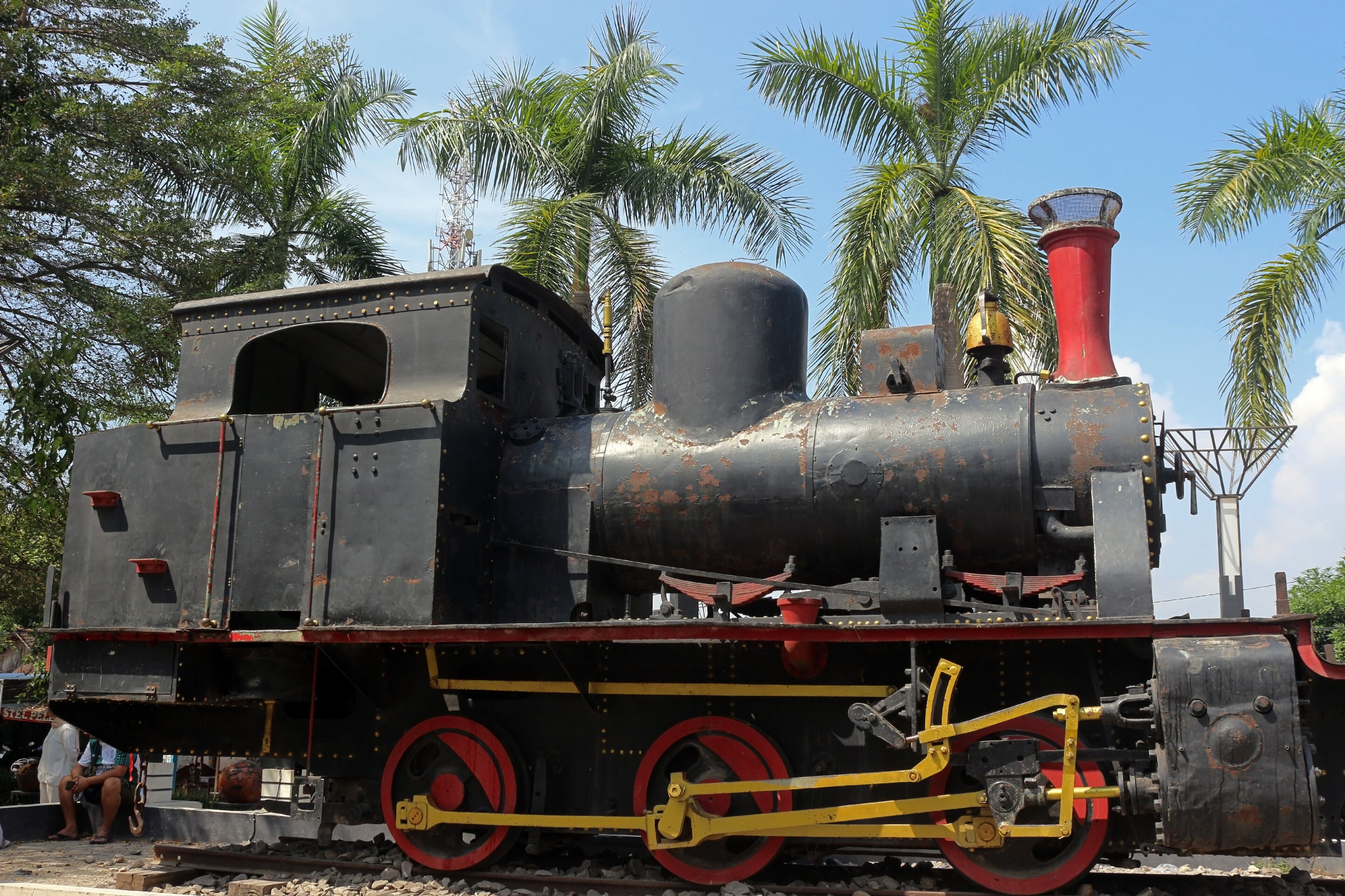 A vintage steam locomotive with red wheels and a red smokestack, parked on tracks with palm trees in the background.