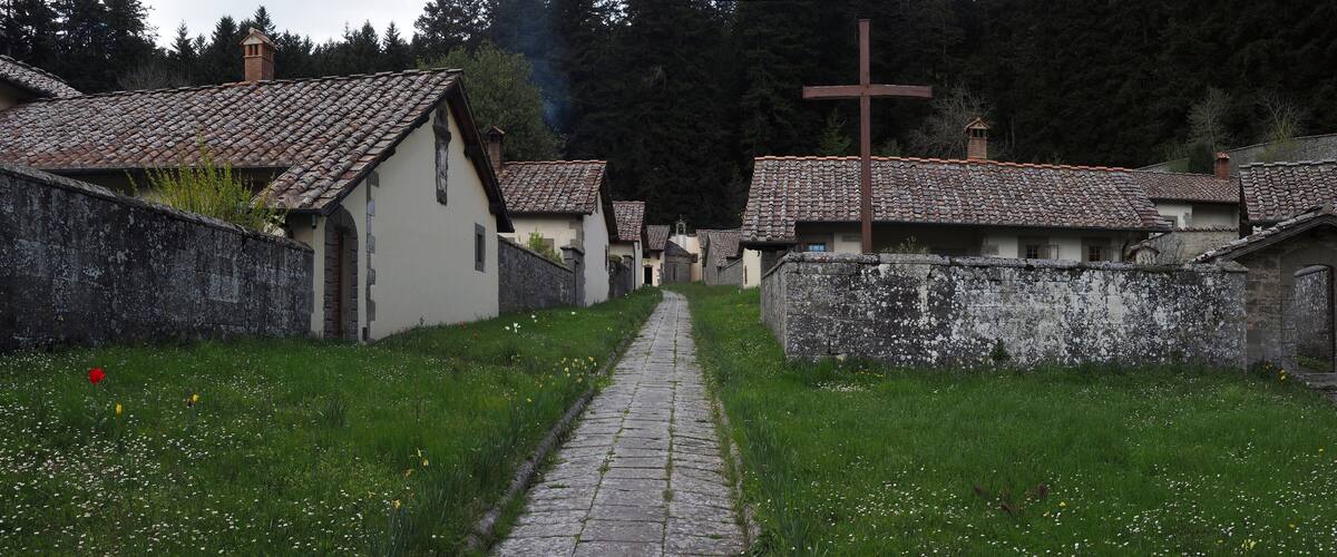 A narrow cobblestone path flanked by traditional stone houses with tiled roofs, set in a lush green landscape with a forested backdrop.