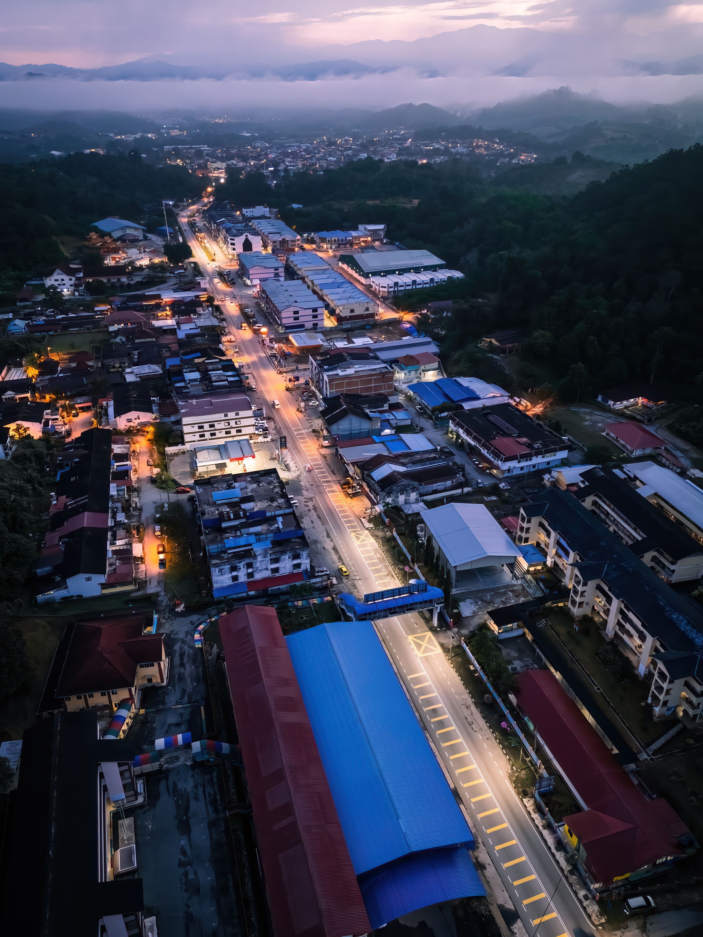 An aerial view of a town at dusk with illuminated streets and buildings, surrounded by mountains and greenery.