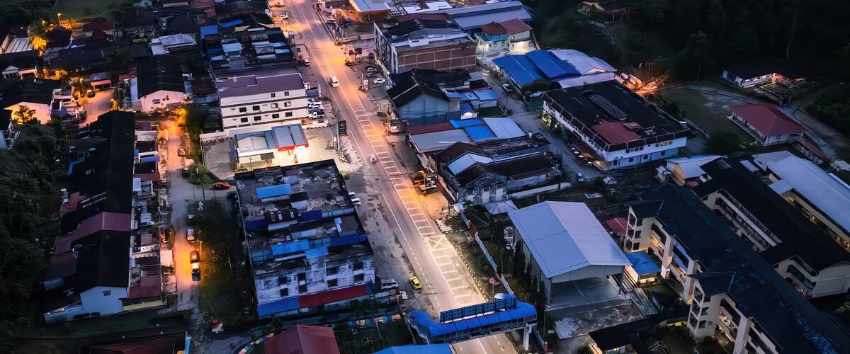 An aerial view of a town at dusk with illuminated streets and buildings, surrounded by mountains and greenery.