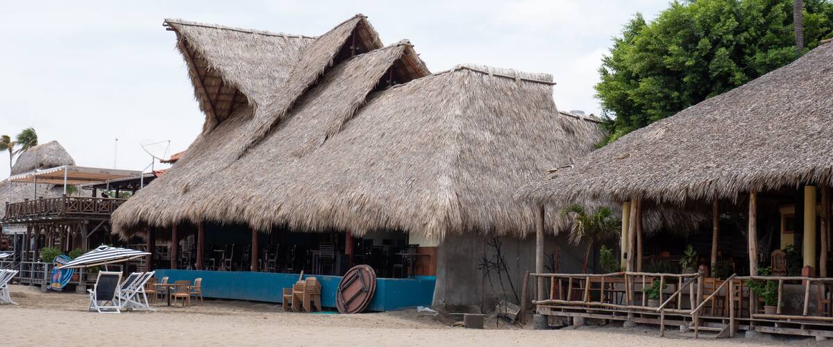 Thatched-roof huts with wooden railings, a sandy beach, and a blue structure with chairs.