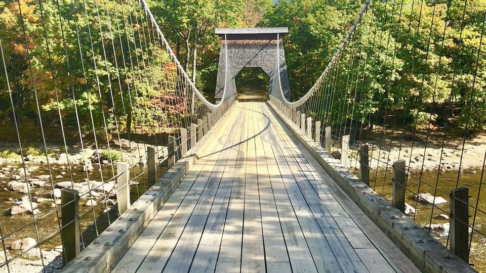 A wooden suspension bridge with a stone archway, surrounded by trees and a river.
