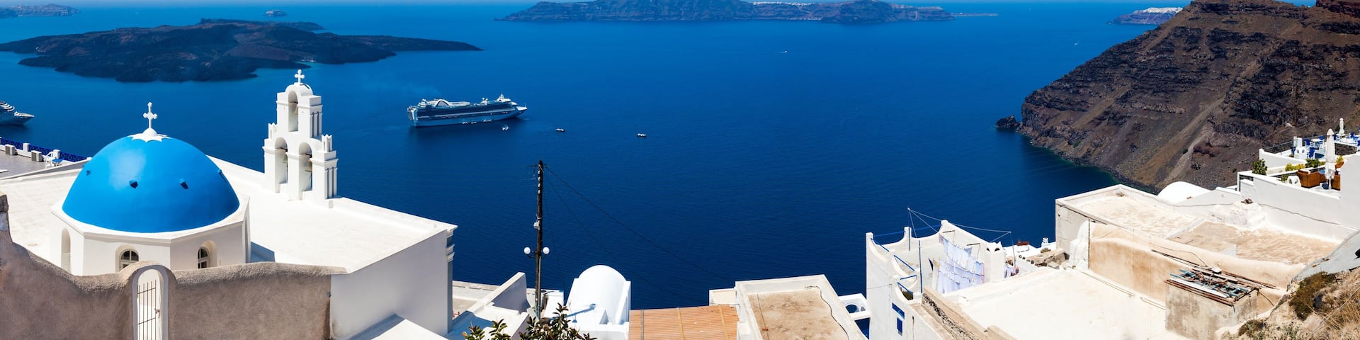 A coastal town with white buildings and blue domes, a large cruise ship, and a clear view of the surrounding islands and sea.